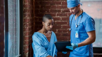 Thoughtful doctor in uniform and gloves showing diagnosis to African American female patient with short hair in blue medical robe in hallway of hospital in daytime