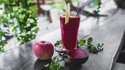 Glass of fresh cold healthy smoothie placed on wooden table on terrace of cafe with whole apple and halved beetroot