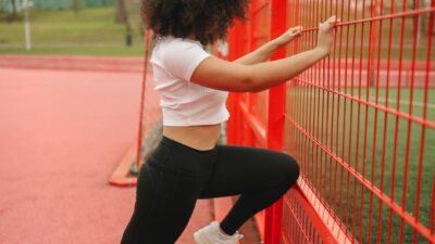 Woman in sportswear stretching against a red metal fence at an outdoor track.