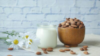 Natural almonds in a wooden bowl with a glass jar of milk and white chamomile flowers on a wood table.