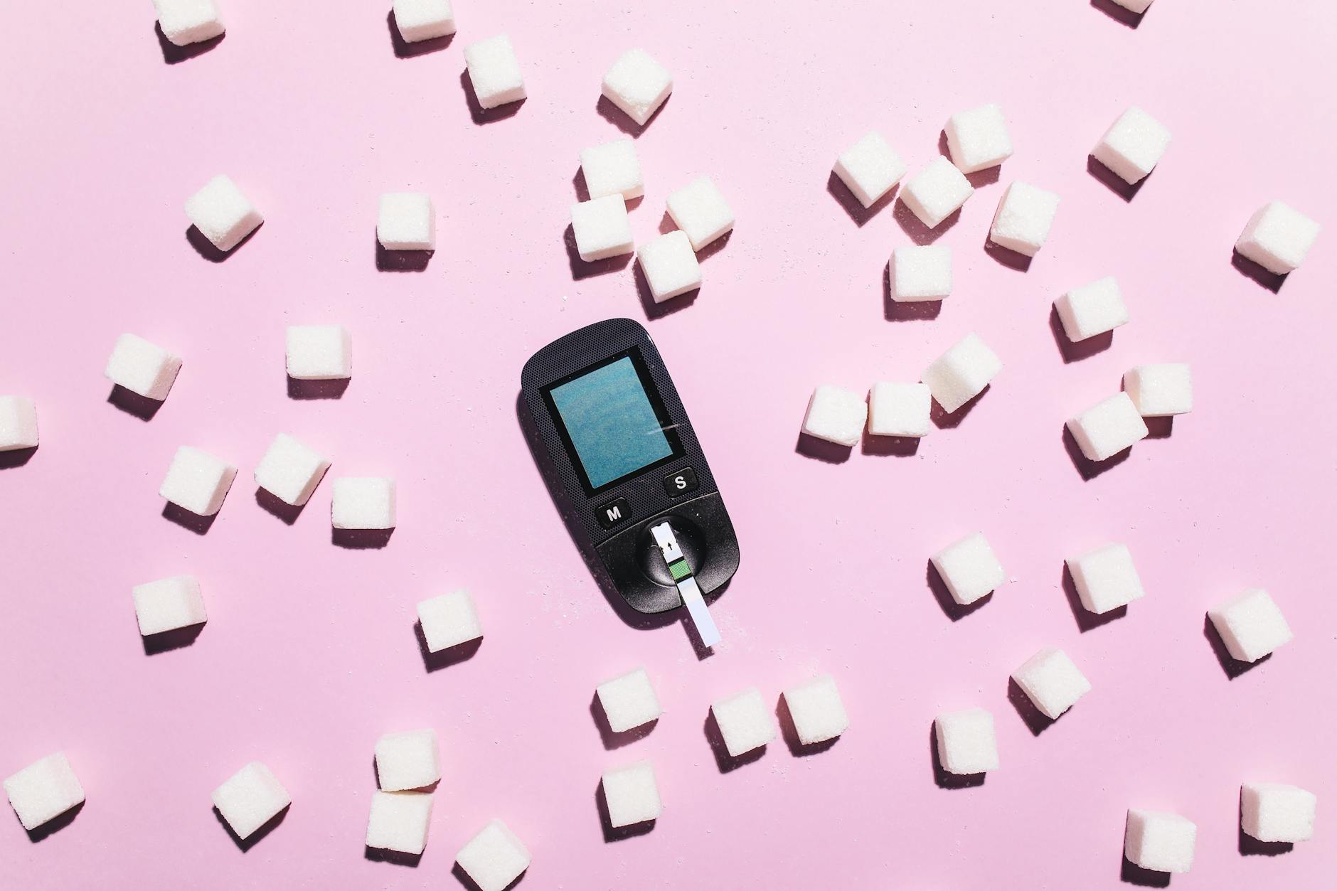 Blood glucose meter surrounded by sugar cubes on a pink background, highlighting diabetes awareness.