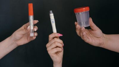 Three hands holding medical tools including a thermometer, lancet, and specimen container.