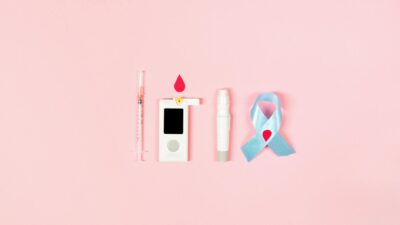 Glucometer, syringe, lancet pen, and ribbon symbolizing diabetes awareness on pink background.