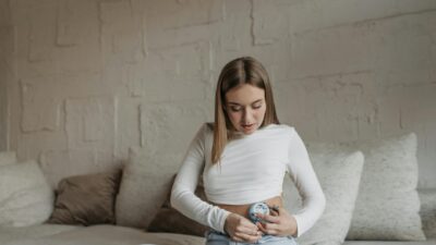 A young woman in white top adjusts her insulin pump while sitting on a sofa indoors.