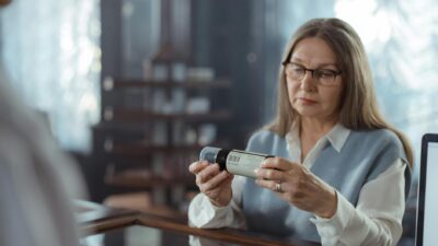 A senior woman with eyeglasses reviews a medicine bottle at a pharmacy counter indoors.