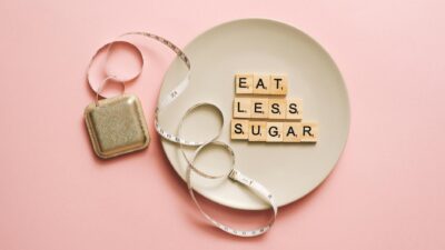 Flat lay of Scrabble tiles saying 'Eat Less Sugar' on a plate with a tape measure on a pink background.
