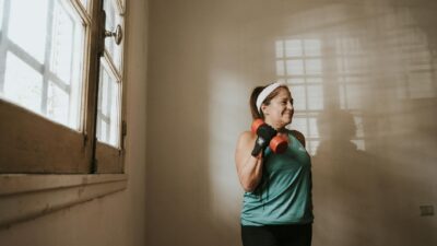 Senior woman in a brightly lit room exercises with dumbbells, promoting fitness at home.