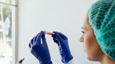A focused lab technician holds a blood sample in a test tube, wearing gloves and a hair net.