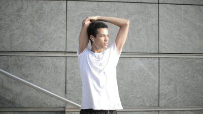 Calm pensive African American sportsman in earphones and sportswear looking away and thinking while stretching arms and listening to music with pleasure on street against gray exterior of contemporary buildings