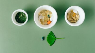 Flatlay of capsules and herbs in ceramic bowls on green surface.