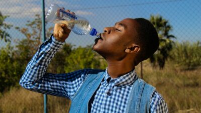 Side profile of a young man enjoying water from a bottle in a sunny outdoor setting.