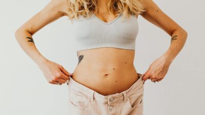A woman displaying weight loss by wearing oversized pants and crop top, showing tattoos.
