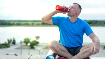 A man in a blue shirt sitting outdoors on an exercise mat, drinking from a water bottle.