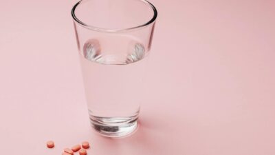 Glass of water with scattered pink pills on a pastel pink background, symbolizing medication or daily supplements.