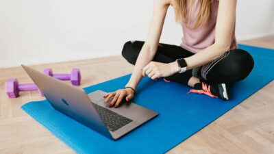 Faceless female in activewear sitting on blue yoga mat on floor and browsing laptop