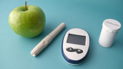Flat lay of a glucose monitor, apple, and insulin pen on a pastel background.