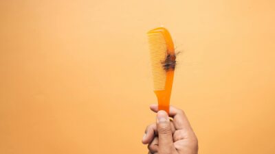 Close-up of hand holding a comb with hair against an orange backdrop, highlighting hair loss concept.