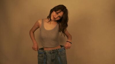Smiling young woman in oversized jeans celebrates weight loss accomplishment.