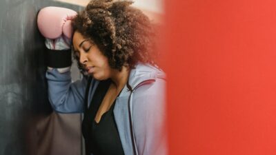 Side view of tired young black female with dark curly hair in sportswear and protective gloves leaning on wall with closed eyes after hard training in gym