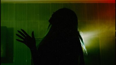 Side view silhouette of female throwing head back and raising hand with spread fingers while standing in dark room against dim green light