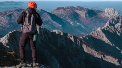 A hiker with a backpack stands on a mountain peak, taking in the panoramic view of rugged mountains.