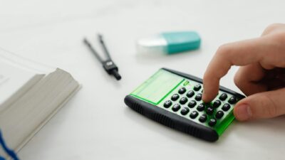 Close-up of a person's hand pressing a calculator on a desk with other stationery items.