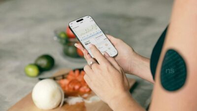 A woman using a smartphone app to track blood sugar levels with fresh vegetables in the background.