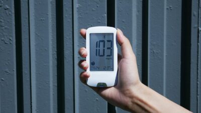 Close-up view of a hand holding a blood glucose meter displaying 103 mg/dL outdoors.