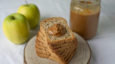 Stack of whole grain bread with peanut butter, accompanied by apples and a jar of smooth peanut butter.