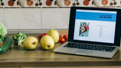 Laptop showing a delivery app beside fresh fruits and vegetables on a kitchen counter.