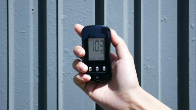 Person holding a glucose meter displaying 103 mg/dL against a textured outdoor background.