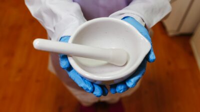Pharmacist holding a white mortar and pestle, wearing blue gloves and lab coat indoors.