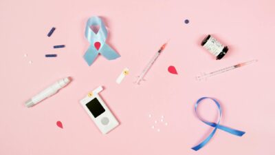 A flat lay image showcasing diabetes awareness items including insulin supplies and a blue ribbon on a pink background.