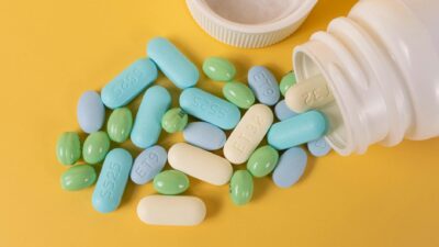 Various colored pills scattered from a white bottle on a yellow surface.