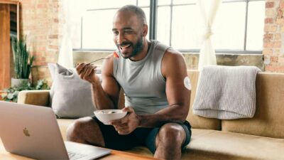 Smiling man eating breakfast and working on a laptop in a cozy home setting.
