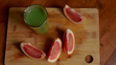 A glass of green juice with fresh grapefruit slices on a wooden cutting board.