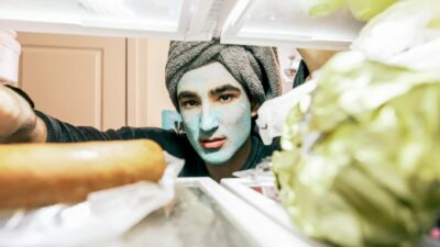 A man with a face mask peers into a refrigerator, wrapped in a towel.
