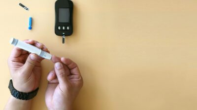 Hands using a glucometer for blood sugar testing on a beige background.