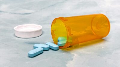 Close-up of blue tablets spilling from an orange prescription bottle on a marble surface.