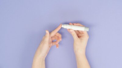 Hands using a lancet pen for blood testing on a light purple background.