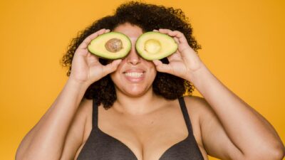 Plump African American woman in bra with curly hair and toothy smile covering eyes with avocado halves