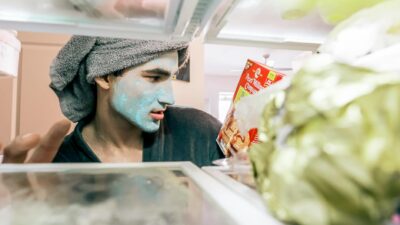 Man with a towel on head and facial mask looking into a refrigerator.