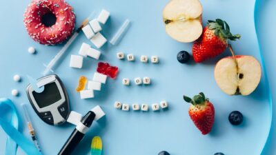 A creative diabetes awareness still life with insulin syringe, fruits, donut, and glucose meter on blue background.