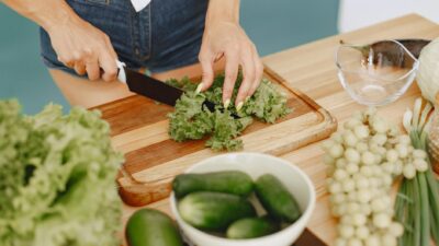 Hands chopping leafy greens on a wooden board with fresh vegetables for a healthy meal preparation.