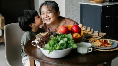 Cute little Asian boy kissing cheek of happy grandmother while sitting at table with fresh healthy apples and salad during lunch in modern kitchen
