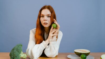 Woman with red hair holding lime surrounded by fresh produce on table.