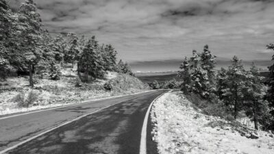 Black and white photo of a winter road cutting through a snowy forest landscape.
