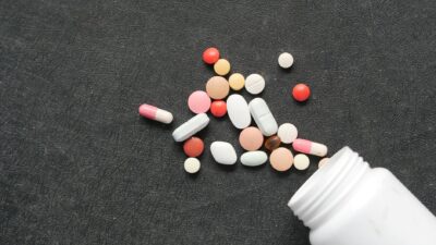 Close-up of diverse pills and capsules spilled from a white bottle on a dark textured surface.