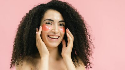 Cheerful woman with curly hair and eye patches smiling against a pink backdrop, promoting skincare.