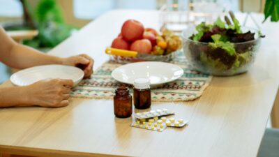 From above of crop anonymous person sitting at table with pile of various medicines and bowls of fresh lettuce salad and ripe fruits in kitchen